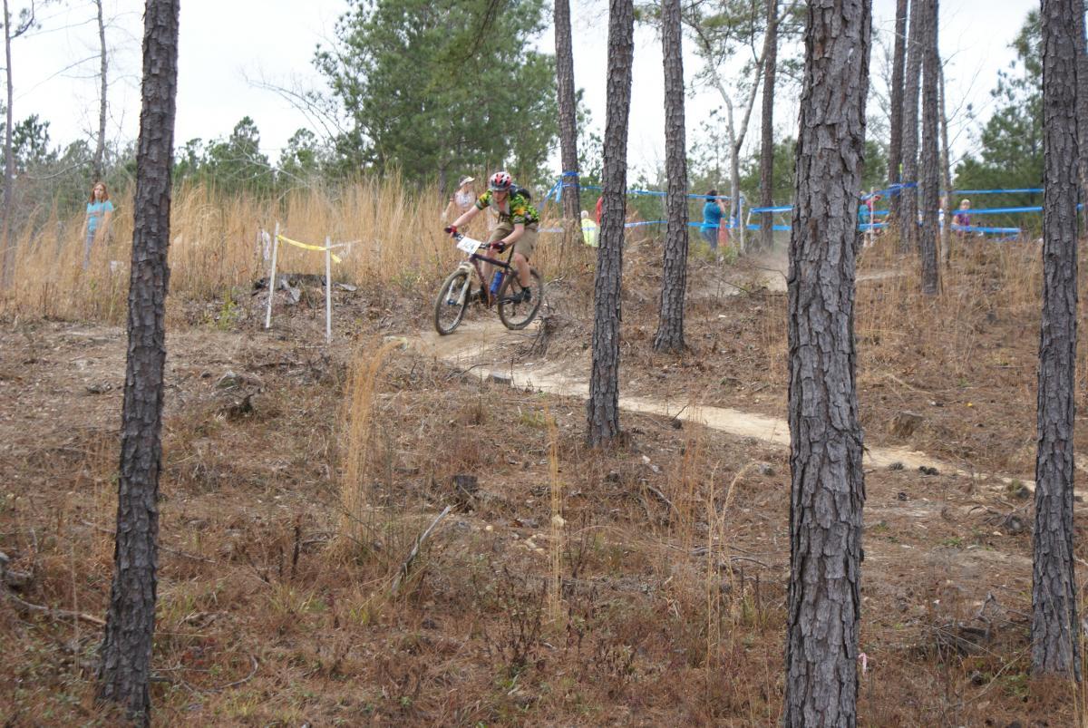 A mountain biker riding along a winding dirt trail through a wooded area. The scene features tall pine trees and sparse underbrush, with a few spectators in the background. The biker is wearing a helmet and a colorful jersey as they navigate the course. Arrowhead Park mountain bike trail.