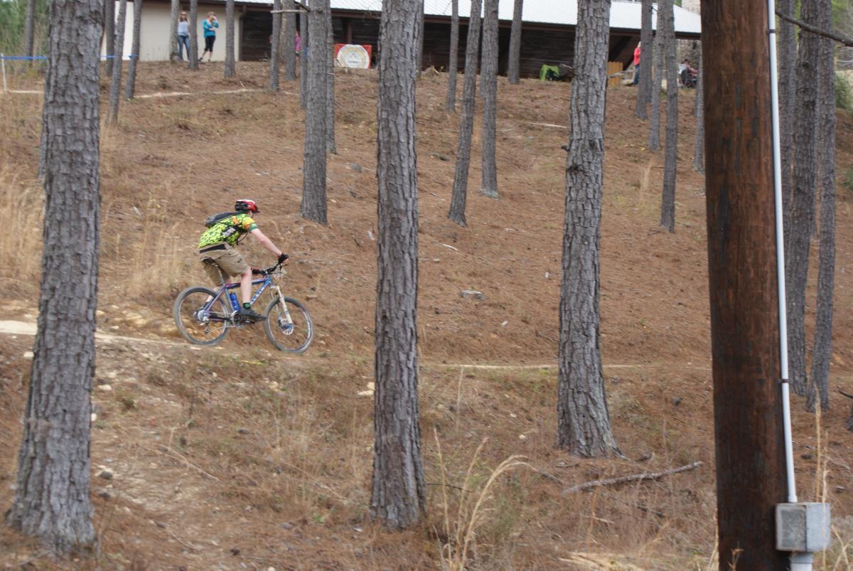 A person riding a mountain bike down a dirt trail through a wooded area with tall pine trees. In the background, a building and a couple of people can be seen. The terrain is sloped, and the cyclist is wearing a helmet and a brightly colored shirt. Arrowhead Park mountain bike trail.
