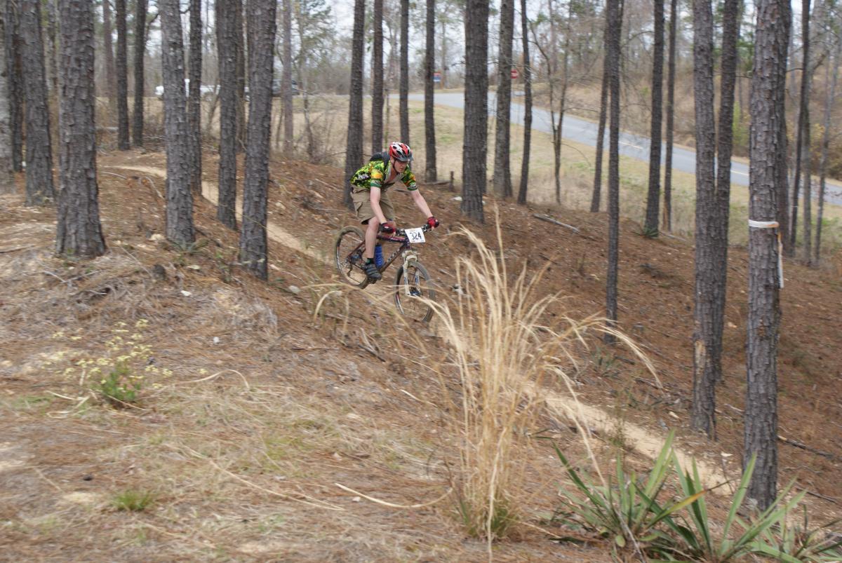A cyclist riding a mountain bike down a dirt trail through a wooded area, surrounded by tall pine trees and sparse grass. The cyclist is wearing a helmet and a colorful jersey, with a race number visible on their bike. In the background, a roadway is visible just beyond the tree line. Arrowhead Park mountain bike trail.