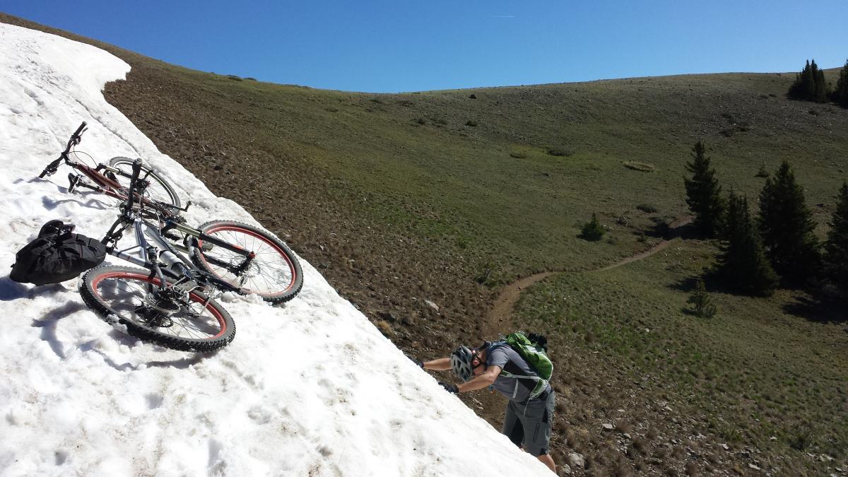 A mountain biker climbing a snowy slope, with two bicycles resting on the snow nearby. The background features a grassy hillside under a clear blue sky, with a winding dirt path visible. Monarch Crest Trail mountain bike trail.