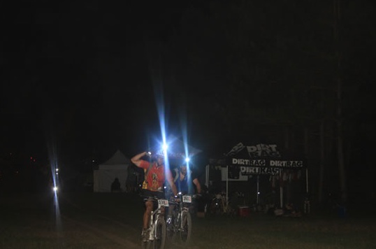 Two mountain bikers ride through a dark field at night, illuminated by their bike headlights. There is a tent in the background with the word "DIRT TAG" visible, along with some additional event equipment and lights. Trees are partially visible on the edges of the scene.