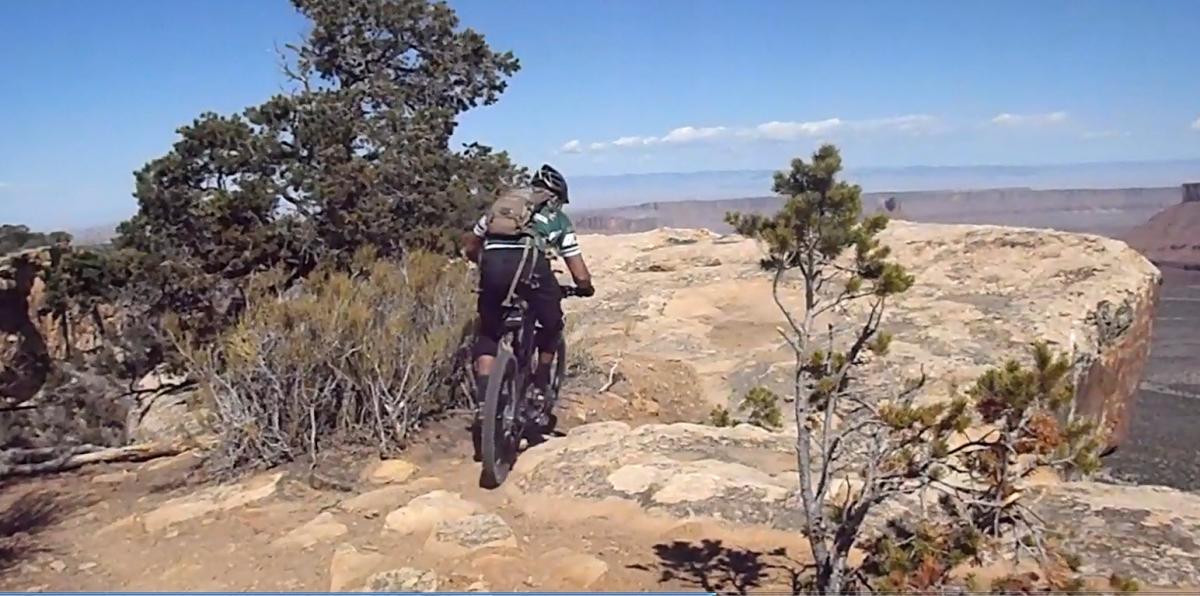 A mountain biker navigates a rocky path along a cliff edge, surrounded by sparse vegetation and towering trees, with a vast landscape and blue sky visible in the background. Porcupine Rim mountain bike trail.