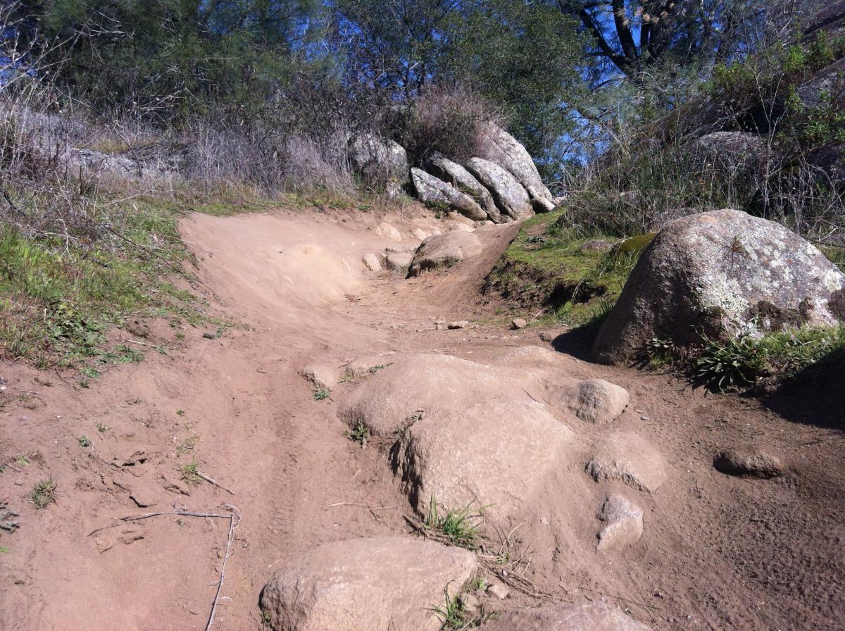 A rugged dirt trail winding through a rocky landscape, lined with boulders and patches of grass, under a clear blue sky. Granite Bay Trail mountain bike trail.