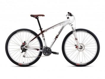 Specialized Rockhopper Disc: A modern mountain bike with a sleek white and red frame, featuring a sturdy front fork, disc brakes, and knobby tires for off-road traction. The bike has a comfortable brown saddle and a standard gear setup, suitable for trail riding.
