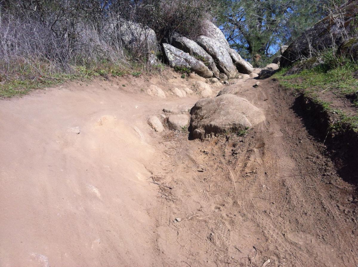 A rocky hiking trail with sandy patches and large boulders on either side, surrounded by sparse vegetation and blue sky above. The path is uneven and shows evidence of use, with tracks leading through the rough terrain. Granite Bay Trail mountain bike trail.