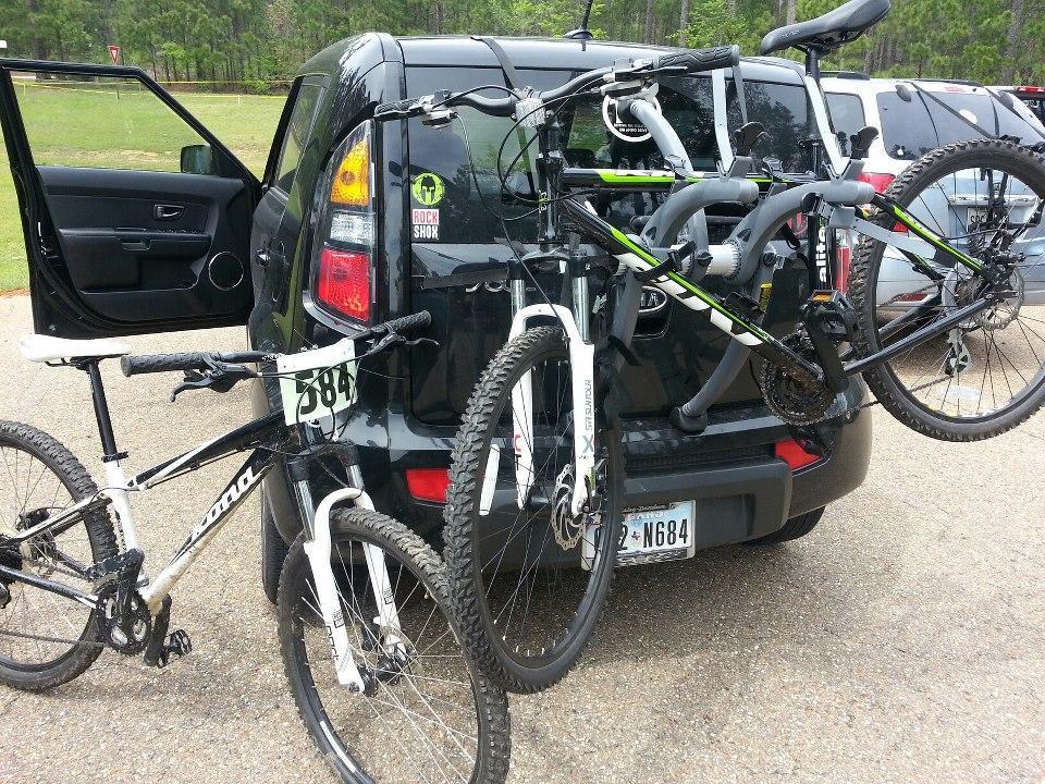 Alt text: A black vehicle with its back doors open, displaying two mountain bikes mounted on a bike rack. One bike is white with black accents, and the other is black with green and white details. The background features a grassy area and other parked cars.