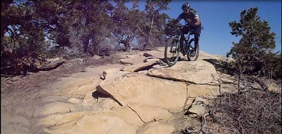 A mountain biker navigating a rocky terrain under clear blue skies, with sparse vegetation in the background. The cyclist is wearing a helmet and riding gear, positioned on a large boulder while maintaining balance on the bike. Porcupine Rim mountain bike trail.
