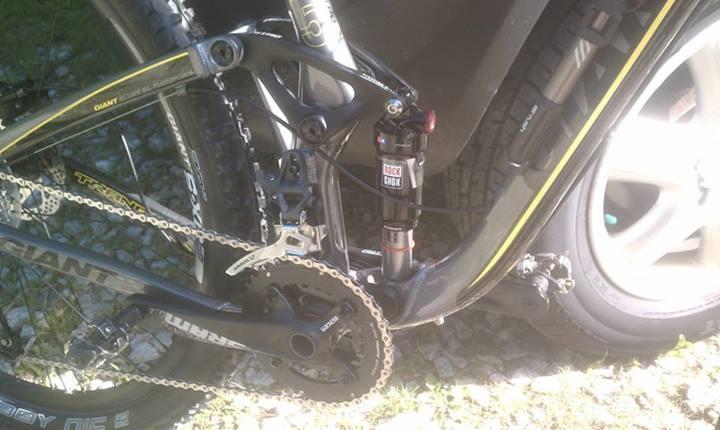 Giant Trance X 29er: Alt text: Close-up view of a bicycle's drivetrain and suspension system, showing the chainring, derailleur, and a shock absorber labeled "ROCKSHOX." The background includes part of a vehicle and some gravel.