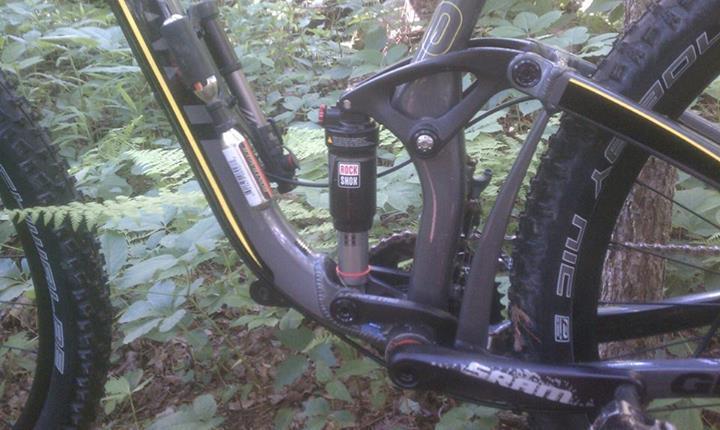 Giant Trance X 29er: Close-up view of a mountain bike's rear suspension system, highlighting the shock absorber labeled "RockShox." The bike frame is black with yellow accents, and is set against a backdrop of greenery and foliage.