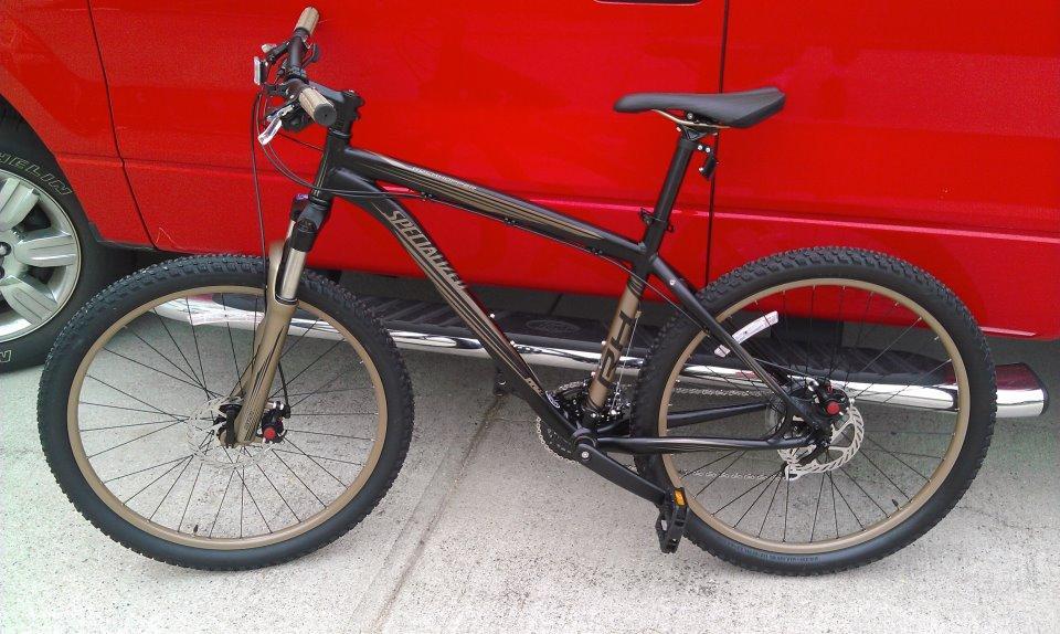 Specialized Rockhopper: A black mountain bike with gold accents parked next to a red vehicle. The bike features thick tires and a sturdy frame, suitable for off-road cycling.