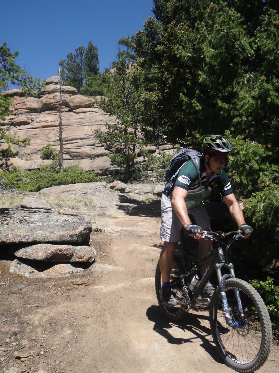 A mountain biker navigating a rocky trail surrounded by dense greenery and rock formations on a sunny day. Staunton State Park mountain bike trail.