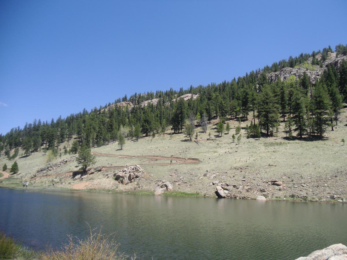 A tranquil lakeside scene featuring a calm body of water bordered by a gravel path and lush greenery. On the opposite shore, a gently sloping hillside is covered with trees, and rocky outcrops are visible. The sky is clear blue with a few fluffy clouds, creating a peaceful outdoor atmosphere. Staunton State Park mountain bike trail.