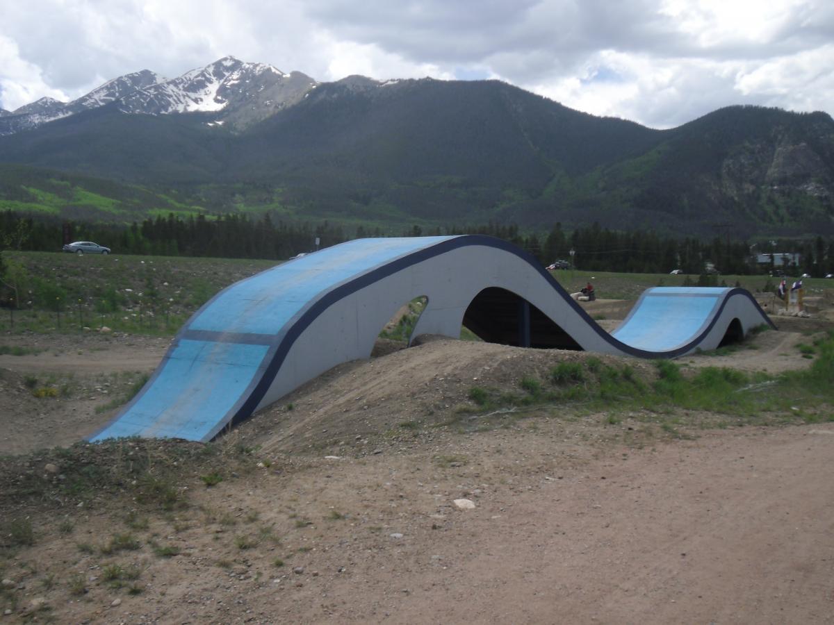 A view of a modern bike pump track featuring curved ramps with blue surfaces, set against a backdrop of green hills and snow-capped mountains under a cloudy sky. Frisco Adventure Park mountain bike trail.