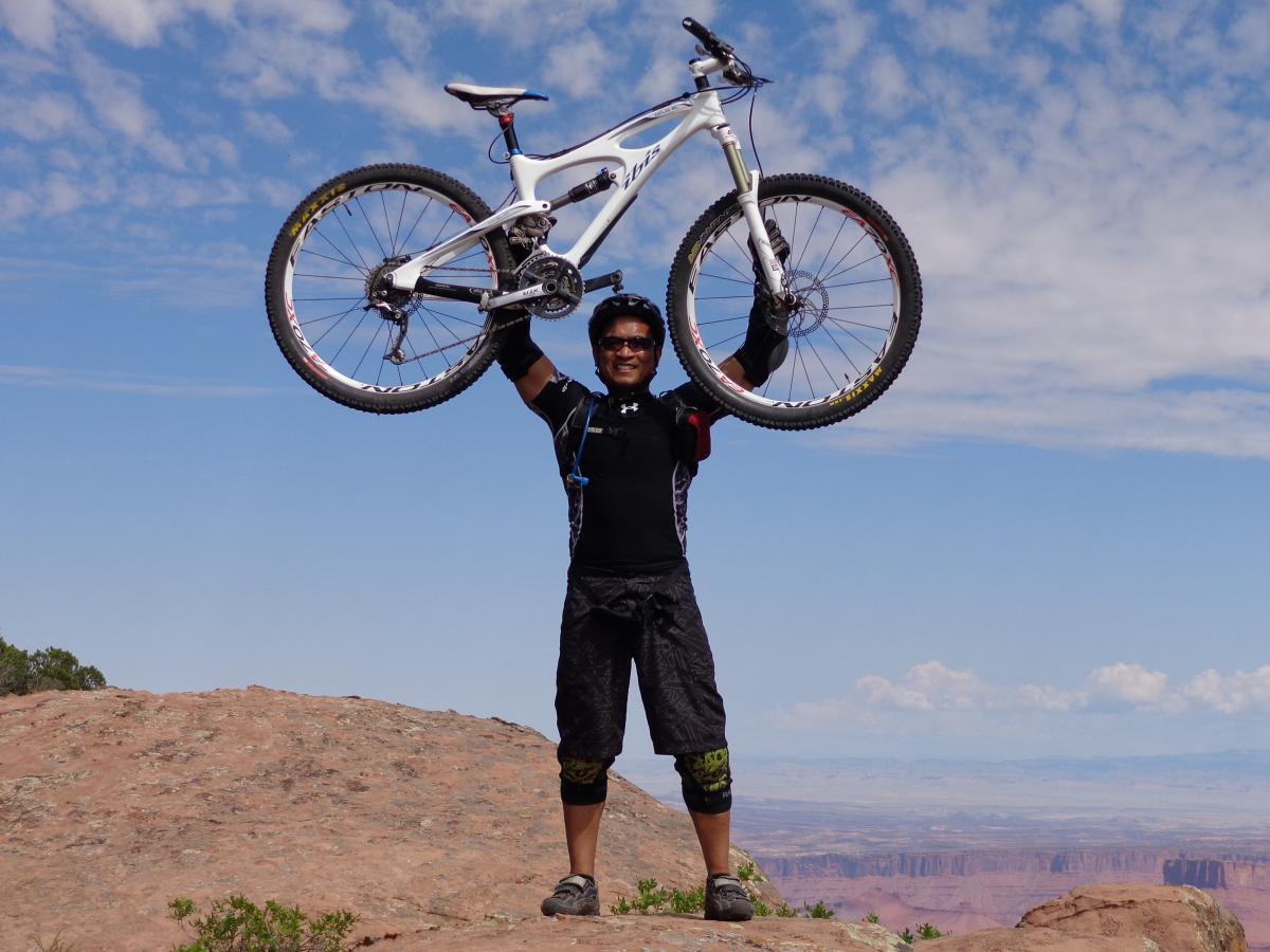 A person dressed in mountain biking gear stands atop a rocky outcrop, triumphantly holding a bicycle above their head. The background features a vast landscape under a blue sky with scattered clouds. The Whole Enchilada mountain bike trail.