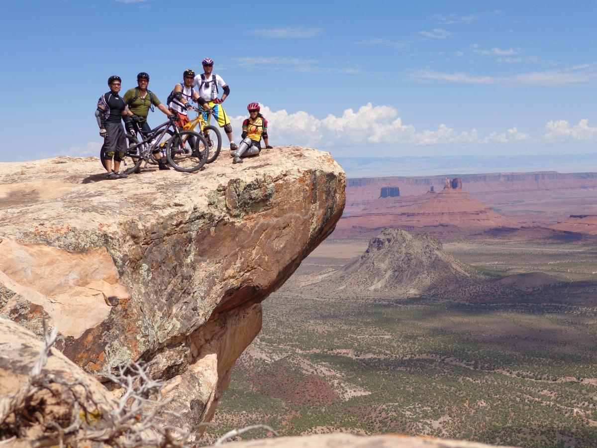 Group of five mountain bikers standing on a rocky outcrop overlooking a vast landscape with distant mesas and hills under a blue sky with some clouds. The Whole Enchilada mountain bike trail.