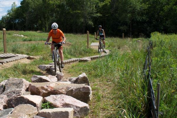 Two mountain bikers navigate a rocky trail in a lush green landscape. One cyclist, wearing an orange shirt and a helmet, rides over a rocky section, while the other cyclist follows on a path lined with tall grasses. Wooden structures are visible in the background, indicating a multi-use trail environment. Lebanon Hills mountain bike trail.