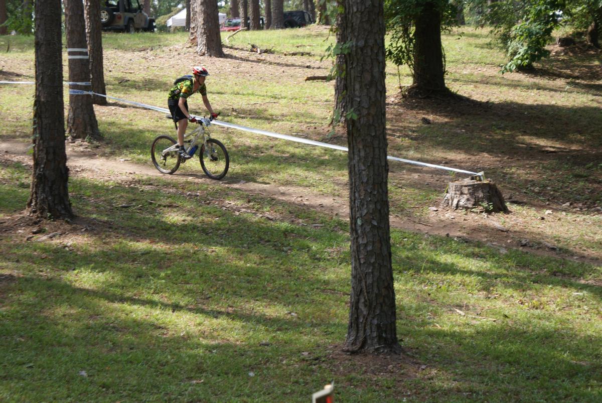 A cyclist rides a mountain bike along a trail in a wooded area, surrounded by trees and grass. The cyclist, wearing a colorful jersey and helmet, is maneuvering past a tree stump, with a course marker visible in the foreground. Flat Rock Park mountain bike trail.
