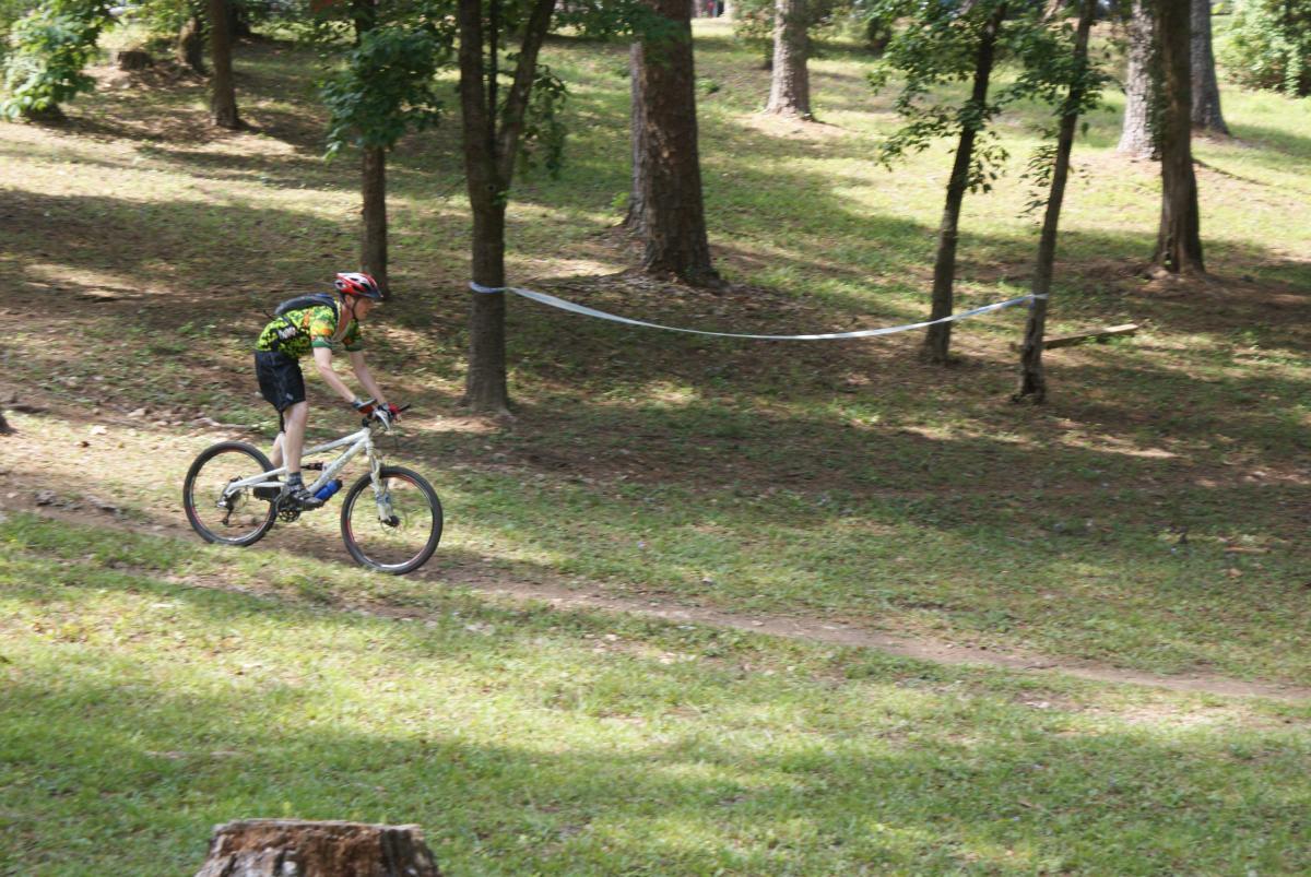 A young boy wearing a colorful cycling jersey and a helmet rides a mountain bike down a grassy path in a wooded area. Trees surround the path, and a ribbon is stretched between two trees, likely marking a course or boundary. The scene is bright and sunny, showcasing a vibrant outdoor setting. Flat Rock Park mountain bike trail.
