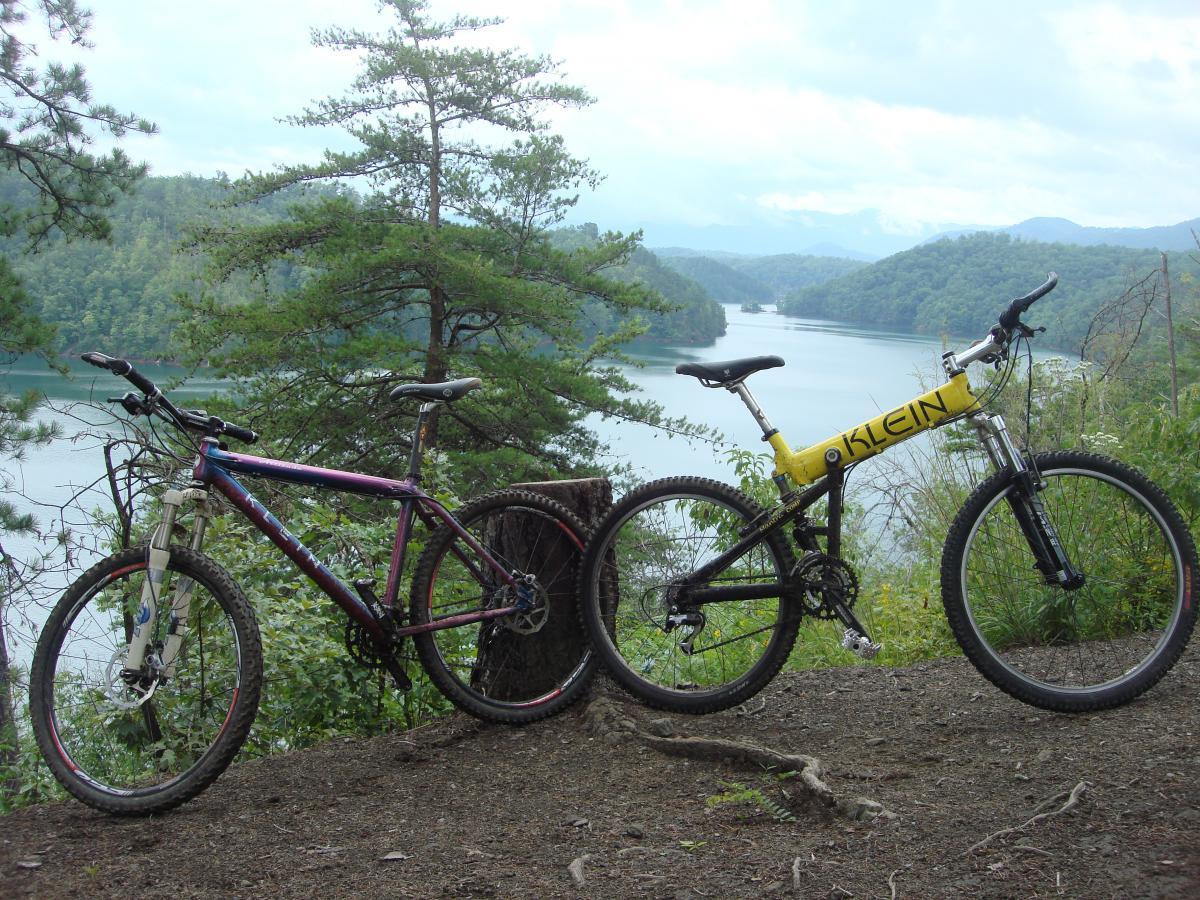 Two mountain bikes are parked on a dirt trail overlooking a serene lake surrounded by green hills and trees. The bike on the left features a purple frame, while the bike on the right has a bright yellow frame with the name "KLEIN" prominently displayed. The scene captures a peaceful outdoor setting perfect for biking enthusiasts. Tsali Recreation Area mountain bike trail.