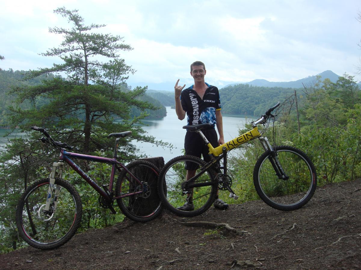 A person standing with two mountain bikes on a trail overlooking a lake surrounded by green hills and trees. The individual is smiling and making a peace sign, dressed in a cycling jersey. Tsali Recreation Area mountain bike trail.