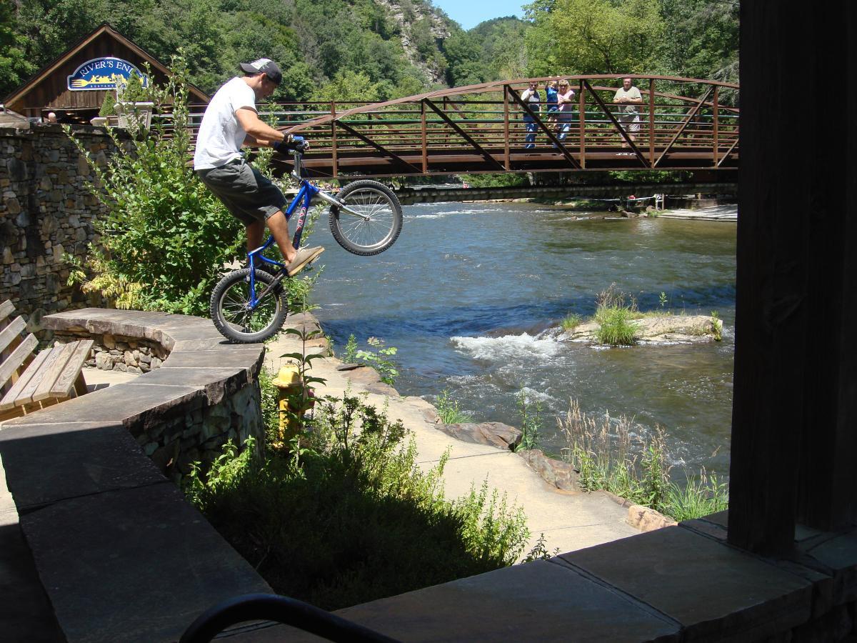 A person performing a stunt on a blue bicycle while balancing on a stone ledge near a river. In the background, a wooden bridge is visible with several onlookers watching the performance. Lush greenery surrounds the scene, and a clear blue sky is present above. Flint Ridge mountain bike trail.