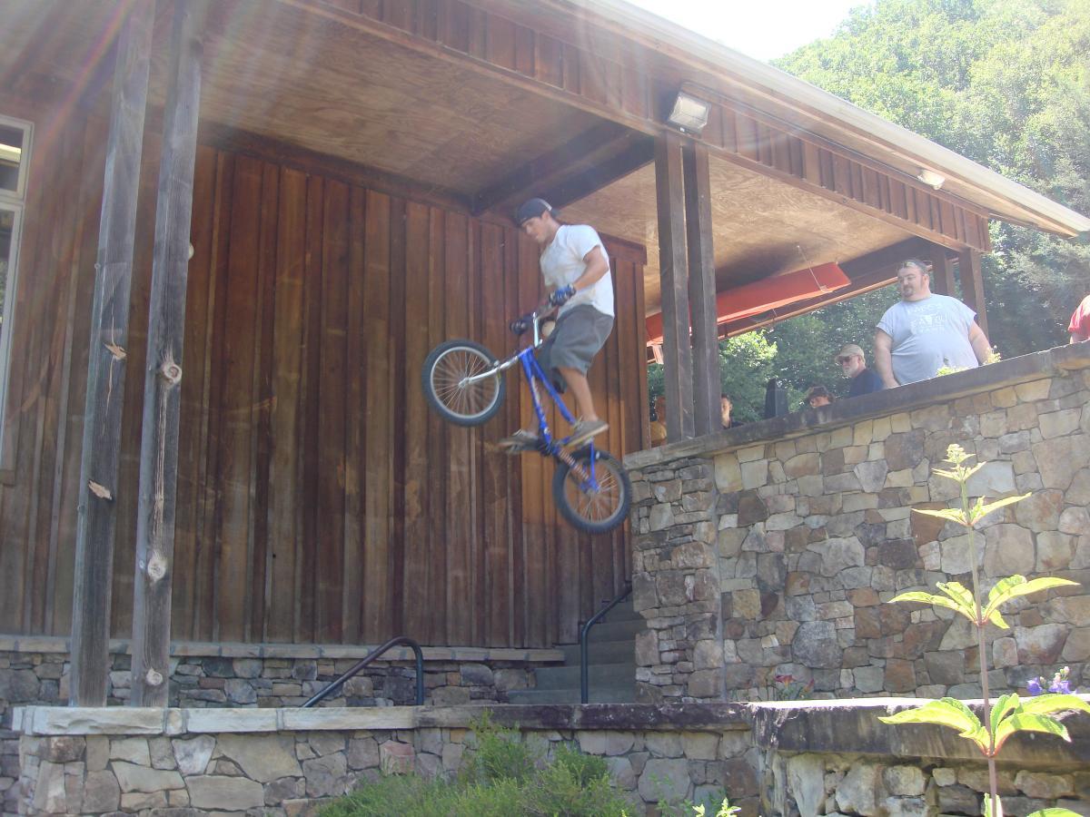 A person riding a blue BMX bike performs a jump off an elevated wooden platform, while a group of onlookers watches from nearby. The scene is set in a sunny outdoor environment with stone walls and greenery in the foreground. Flint Ridge mountain bike trail.