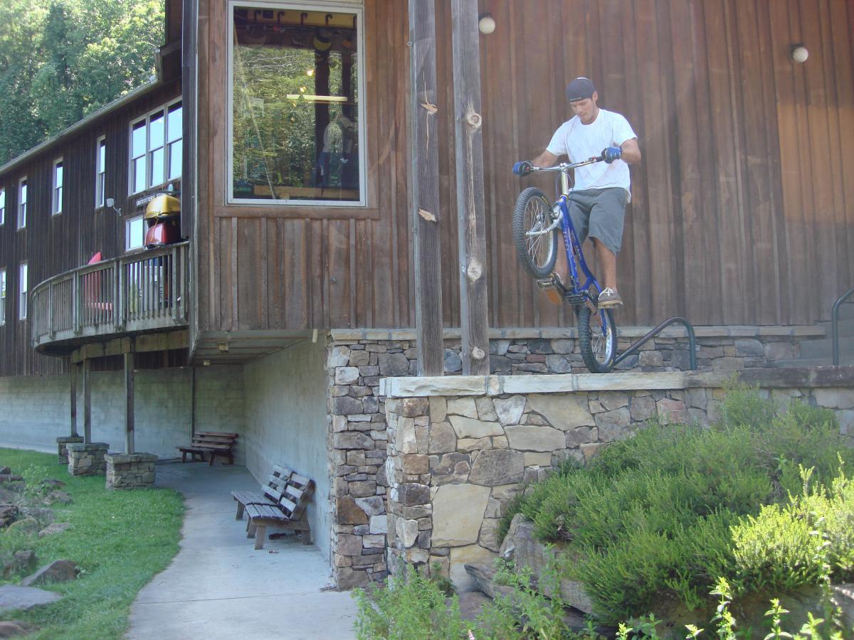 A cyclist performing a trick on a blue bicycle, balancing on a railing next to a wooden building. The setting features a grassy area with benches and stone landscaping, under a sunny sky. Flint Ridge mountain bike trail.