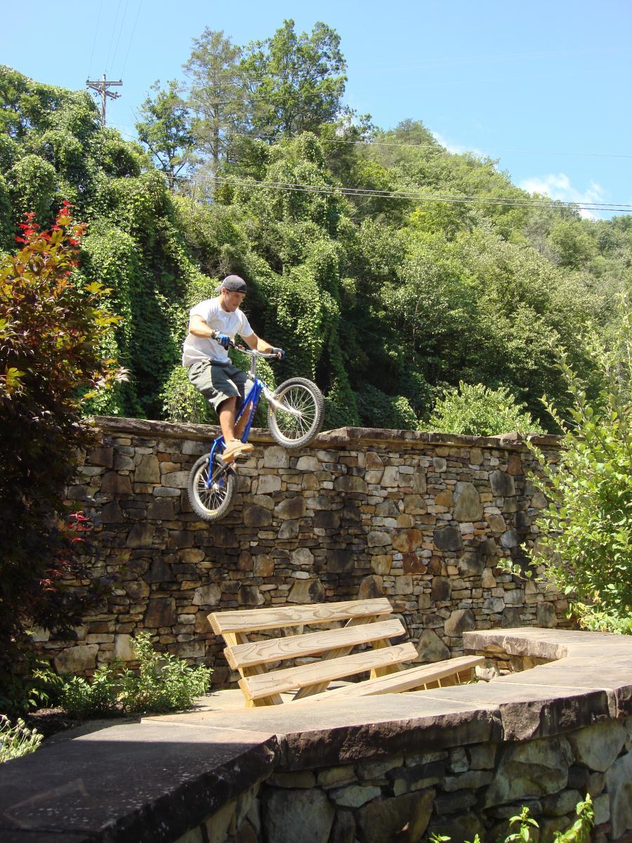 A person on a bicycle performing a jump off a stone wall, with a wooden bench visible below. The background features lush green trees and a clear blue sky. Flint Ridge mountain bike trail.