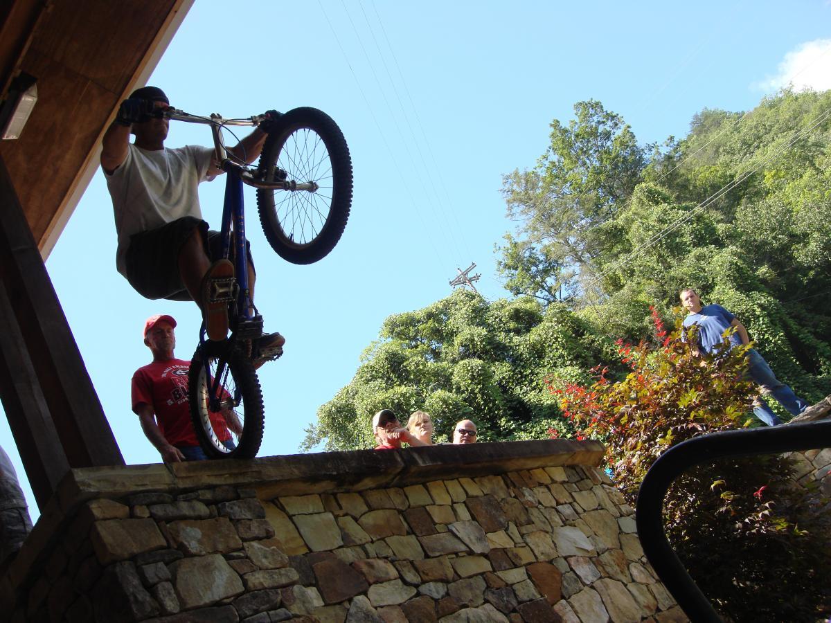 A cyclist performs a trick, riding a bicycle on one wheel while balancing on the edge of a stone wall. Spectators watch the stunt from a distance, surrounded by green trees and clear blue skies. Flint Ridge mountain bike trail.