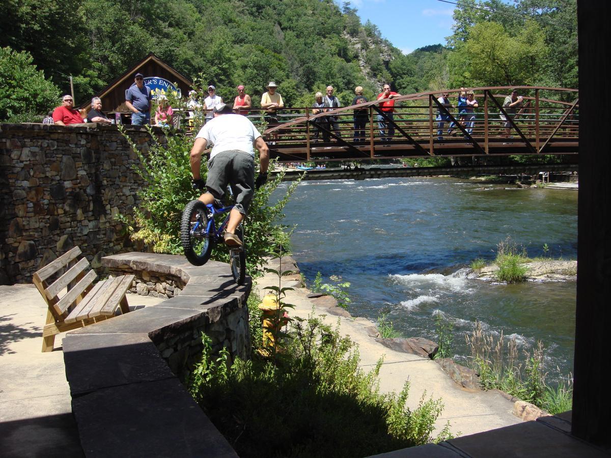A person performing a stunt on a bicycle, jumping over a stone wall near a river, while a crowd of spectators watches from a nearby bridge. Lush greenery surrounds the scene, and the sun is shining brightly. Flint Ridge mountain bike trail.