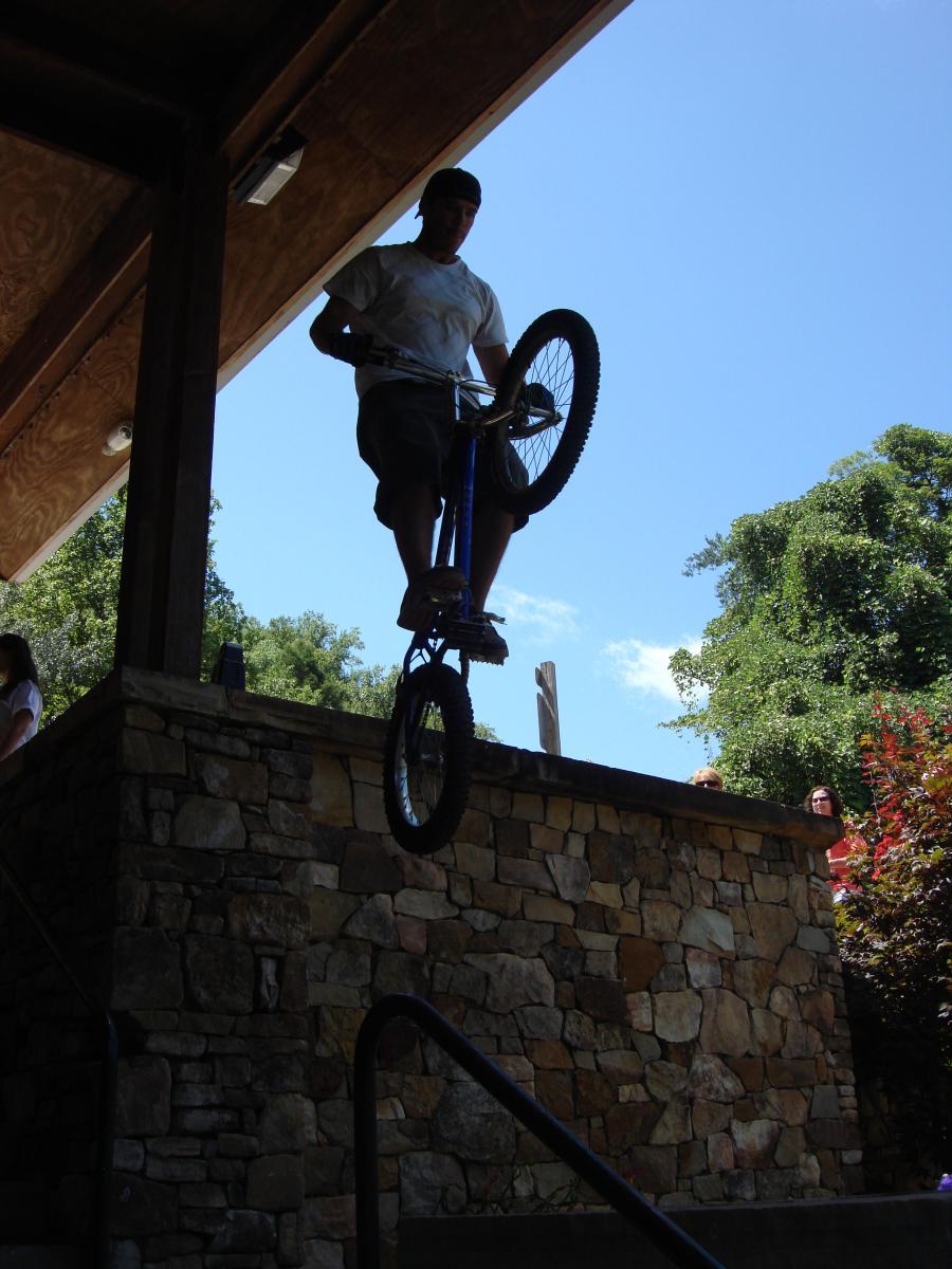 A mountain biker performing a wheelie on a bike while positioned on a raised stone platform. The sky is clear and blue, and a few spectators can be seen watching the stunt from nearby steps and foliage. Flint Ridge mountain bike trail.