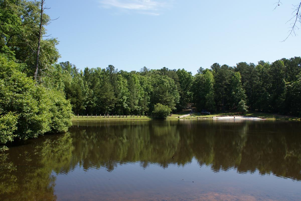 A tranquil lake surrounded by vibrant green trees, reflecting the blue sky above. A small sandy area is visible on the right, with a few people in the distance, and a parked car nearby. Dauset Trails Nature Center mountain bike trail.