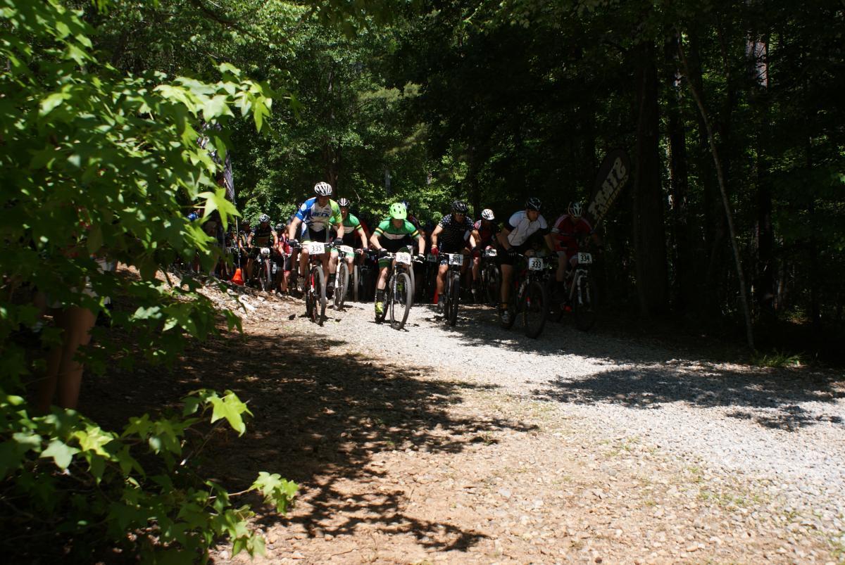 A group of mountain bikers is racing along a gravel path in a wooded area. The riders, wearing colorful jerseys and helmets, are in a competitive start position, with trees and foliage visible on either side of the trail. Dauset Trails Nature Center mountain bike trail.