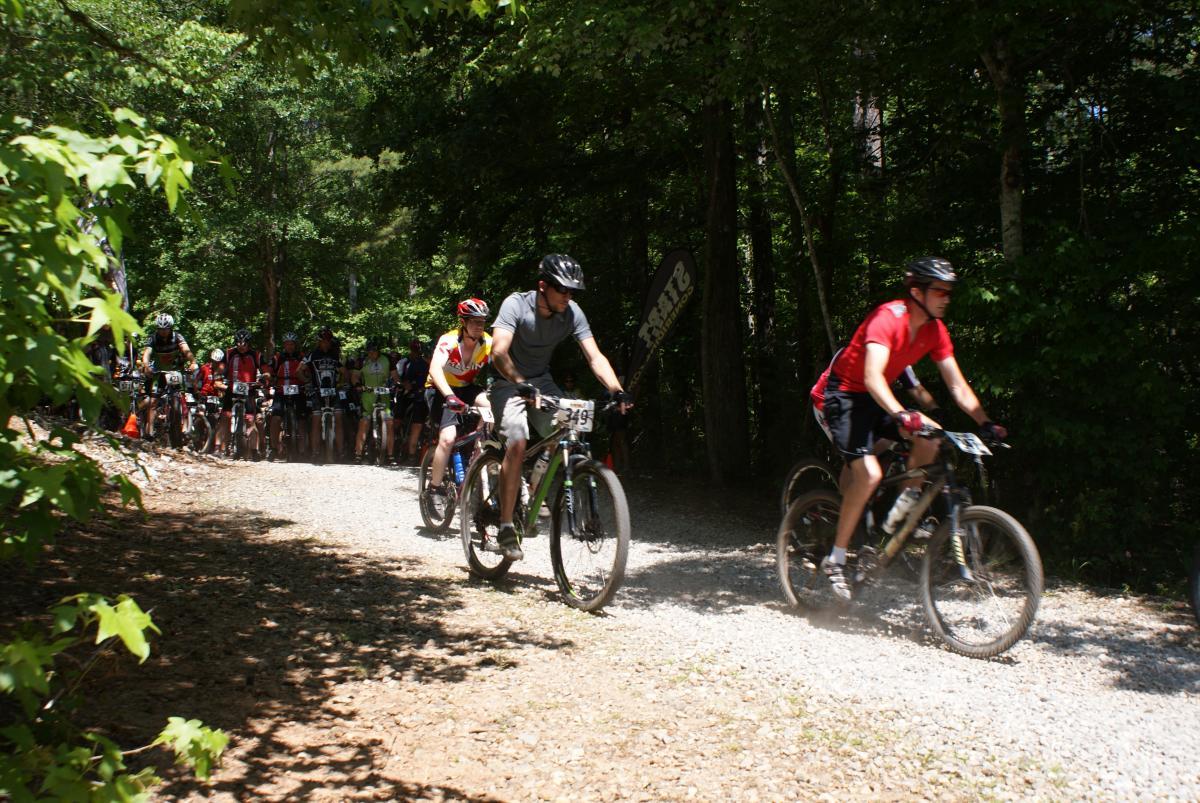 A group of mountain bikers on a gravel trail surrounded by trees, with several cyclists racing ahead and spectators in the background. Some riders are wearing colorful jerseys, and flags can be seen indicating the event. The scene captures the excitement of a biking competition in a natural setting. Dauset Trails Nature Center mountain bike trail.