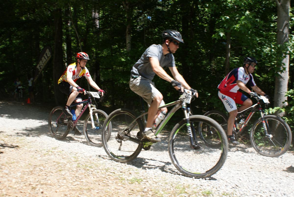 Three mountain bikers riding on a rocky trail through a wooded area. The first rider wears a colorful jersey and is in the lead, followed closely by two others. The scene captures the intensity of the biking competition, with trees and sunlight in the background. Dauset Trails Nature Center mountain bike trail.