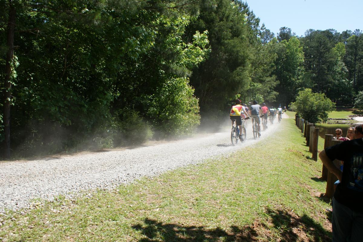 A group of cyclists riding on a gravel path surrounded by trees, with dust being kicked up behind them. In the foreground, a few spectators watch the cyclists from a grassy area next to the path. Dauset Trails Nature Center mountain bike trail.