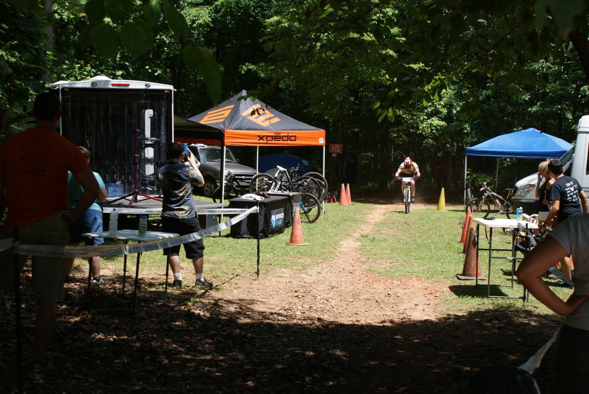 A mountain biker speeds down a dirt path during a competitive cycling event. In the background, spectators watch from behind a booth with bikes and equipment, while colorful tents provide shade in a wooded area. Several individuals are seen capturing the moment with cameras. Dauset Trails Nature Center mountain bike trail.