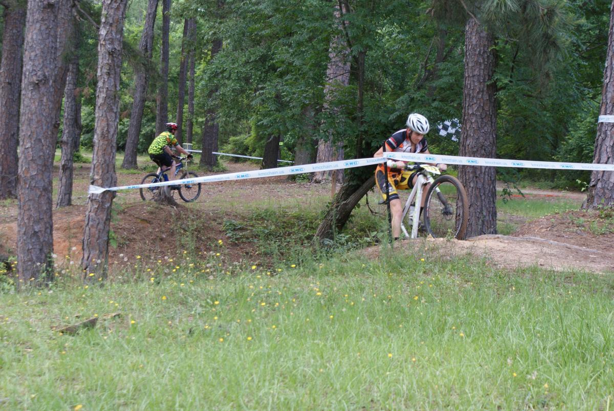 Two mountain bikers navigating a forested trail. One rider in a green jersey is seen cycling around a curve, while the second rider, dressed in a striped and orange shirt, is maneuvering over a small rise. Tall trees and greenery surround the dirt path, with a race ribbon visible in the background. Flat Rock Park mountain bike trail.