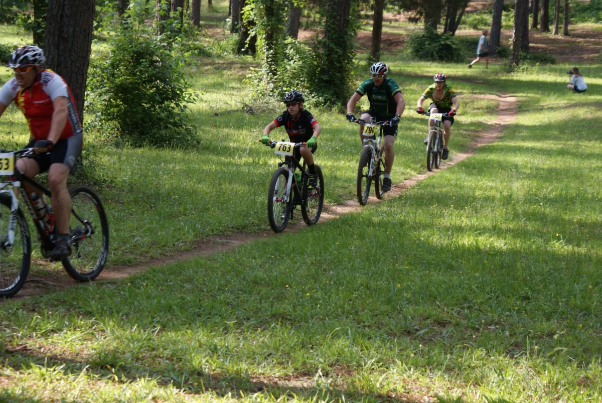 A group of four mountain bikers navigating a grassy trail in a wooded area during a biking event. The riders are wearing helmets and numbered bibs, with a mix of colorful jerseys. Lush greenery surrounds the path, and additional spectators can be seen in the background. Flat Rock Park mountain bike trail.