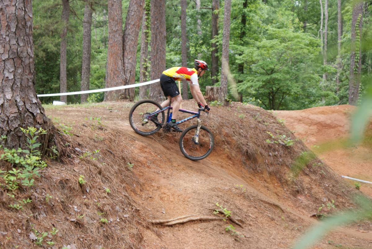 A mountain biker navigating a dirt trail with a slight incline, surrounded by tall trees and green foliage. The rider is wearing a helmet and colorful cycling gear, positioned on a blue mountain bike. A white racing tape is visible in the background, indicating a course boundary. Flat Rock Park mountain bike trail.