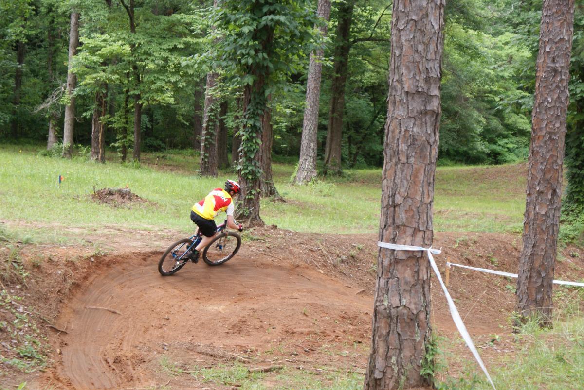 A cyclist in a red and yellow jersey rides on a dirt trail through a forested area, navigating a banked turn surrounded by tall trees and greenery. Flat Rock Park mountain bike trail.