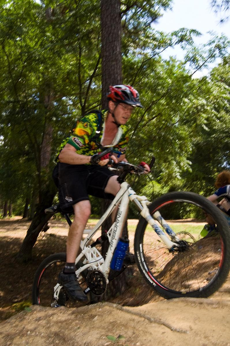 A mountain biker in a colorful jersey and helmet rides over a dirt mound in a forested area, with trees in the background. The bike is white with visible suspension components, and a blue water bottle is attached to the frame. Another cyclist is seen in the background. The image captures the dynamic movement and energy of mountain biking. Flat Rock Park mountain bike trail.