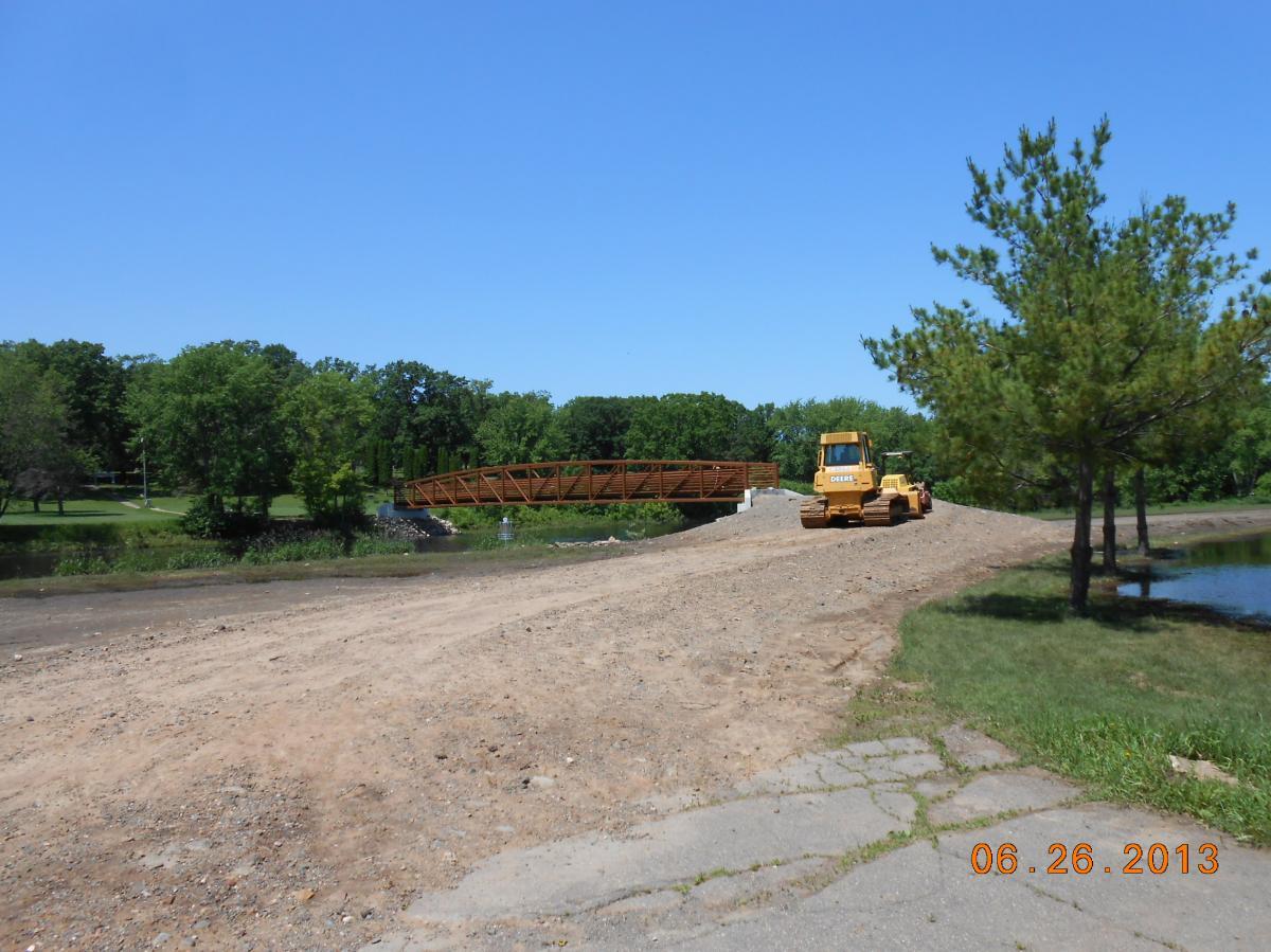 A construction site featuring a yellow bulldozer parked near a recently built wooden bridge over a body of water. The scene is set against a backdrop of trees and a clear blue sky. The area is mostly bare earth and gravel, indicating ongoing construction or landscaping work. The date in the bottom right corner reads June 26, 2013. Milaca trails mountain bike trail.