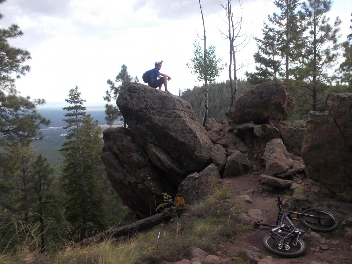 A person sitting on a large rock formation overlooking a scenic view of trees and distant hills, with a mountain bike lying on the ground nearby. The sky is partly cloudy, adding to the outdoor adventure atmosphere. Schultz Creek mountain bike trail.
