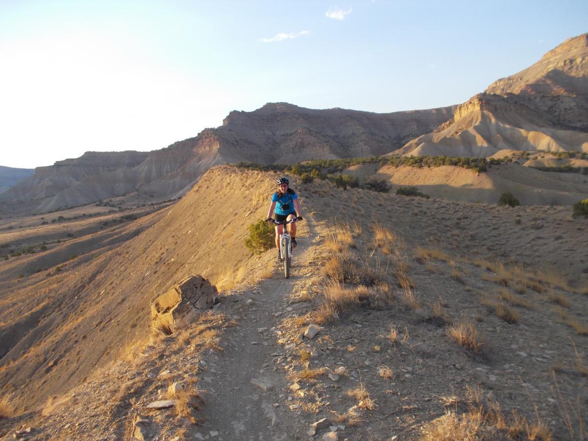 A mountain biker riding along a narrow dirt trail on a rocky hillside, surrounded by rugged terrain and distant mountains under a clear sky. The landscape features dry grass and sparse vegetation, with warm sunlight illuminating the scene. 18 Road Trails / North Fruita Desert mountain bike trail.