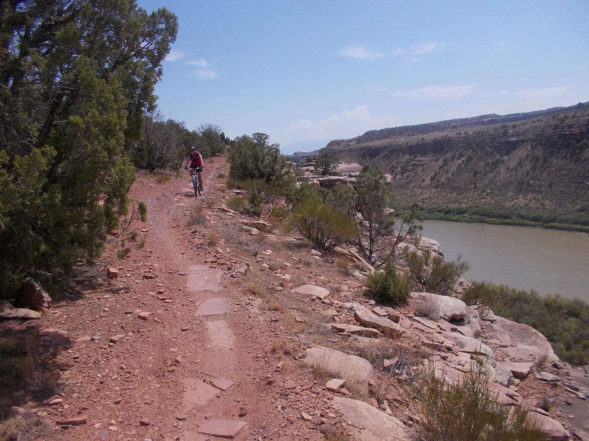 A mountain biker rides along a narrow, rocky trail on a hillside overlooking a river. The surrounding landscape features sparse vegetation, including shrubs and trees, against a backdrop of rugged cliffs and a blue sky with scattered clouds. Mary's Loop / Horsethief Bench mountain bike trail.