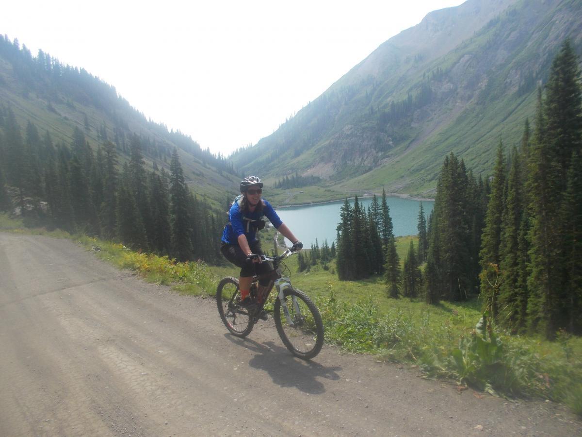 A mountain biker rides along a dirt path surrounded by lush green hills and tall pine trees, with a serene blue lake visible in the background. The scene is illuminated by soft natural light, creating a picturesque outdoor atmosphere. Trail 401 mountain bike trail.