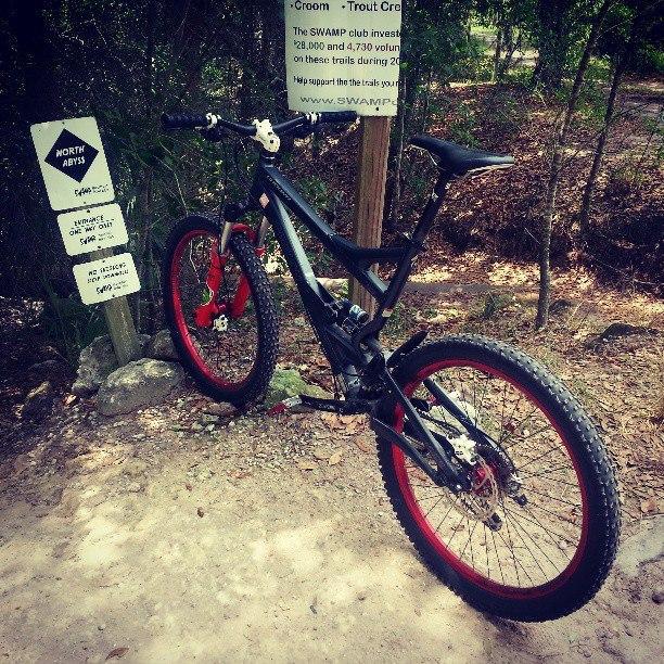 Specialized Enduro Elite: A black mountain bike with red accents leans against a wooden signpost surrounded by trees and dirt trails. The sign indicates trail information, with one sign reading "NORTH ARROWS" and additional text highlighting trail management efforts. Sunlight filters through the foliage, creating a natural outdoor setting.