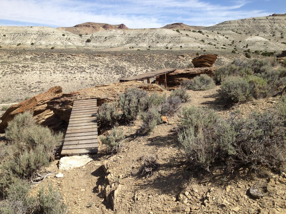 A wooden walkway extends over rocky terrain, leading through sparse vegetation and sagebrush. The landscape features rolling hills in the background with a clear blue sky and scattered clouds. Pick Your Poison mountain bike trail.