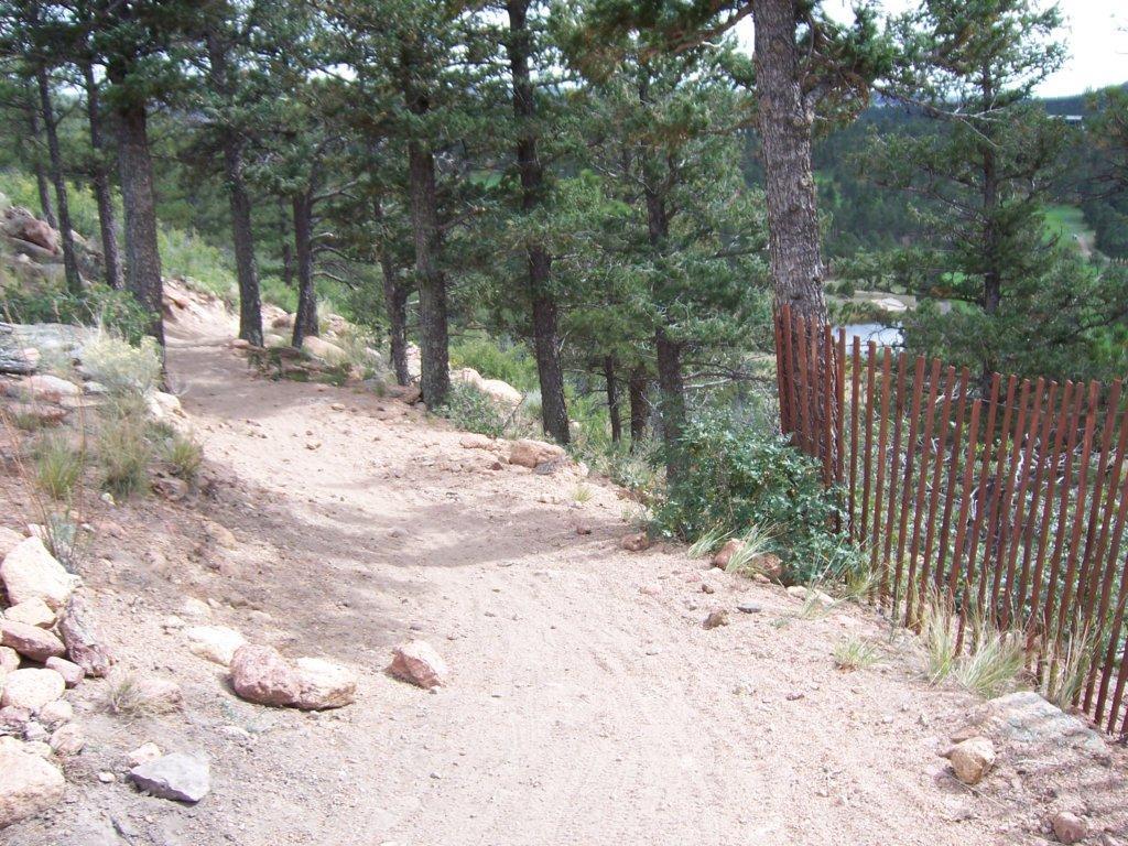 A winding dirt trail lined with tall pine trees, leading through a natural landscape. The path is surrounded by rocky terrain and a wooden fence in the background, with a glimpse of a body of water visible through the trees. Falcon Trail mountain bike trail.